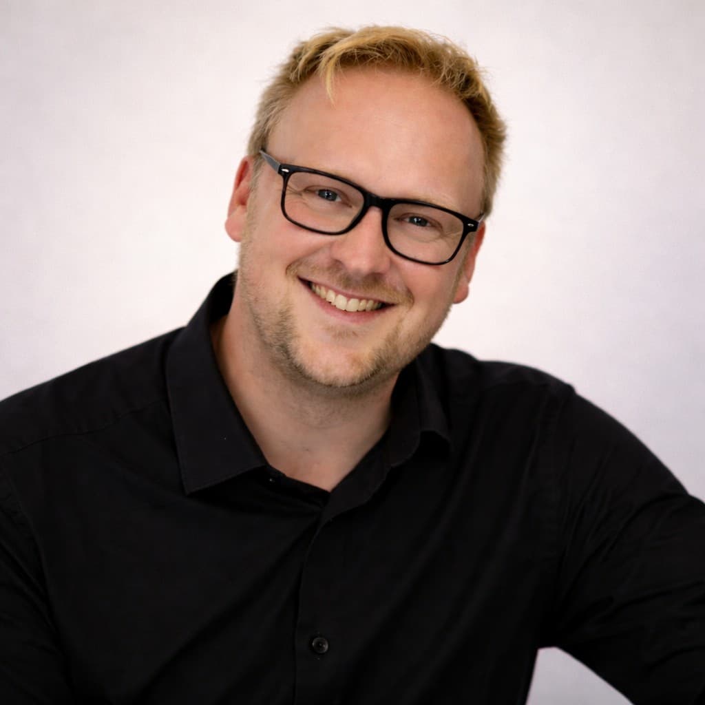 Portrét Lukáše Sádeckého s úsměvem, v černých brýlích a černé košili, neutrální světlé pozadí.